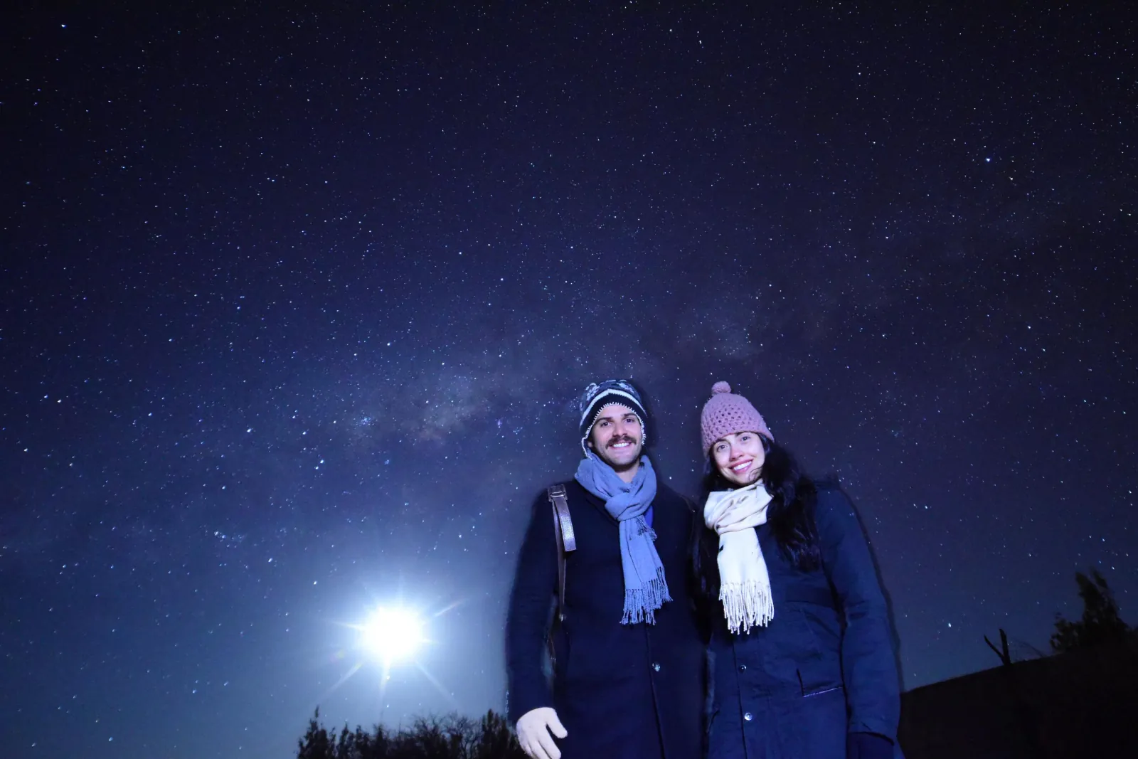 A couple in beanies and scarves poses under the Milky Way as a bright moon shines low beside them