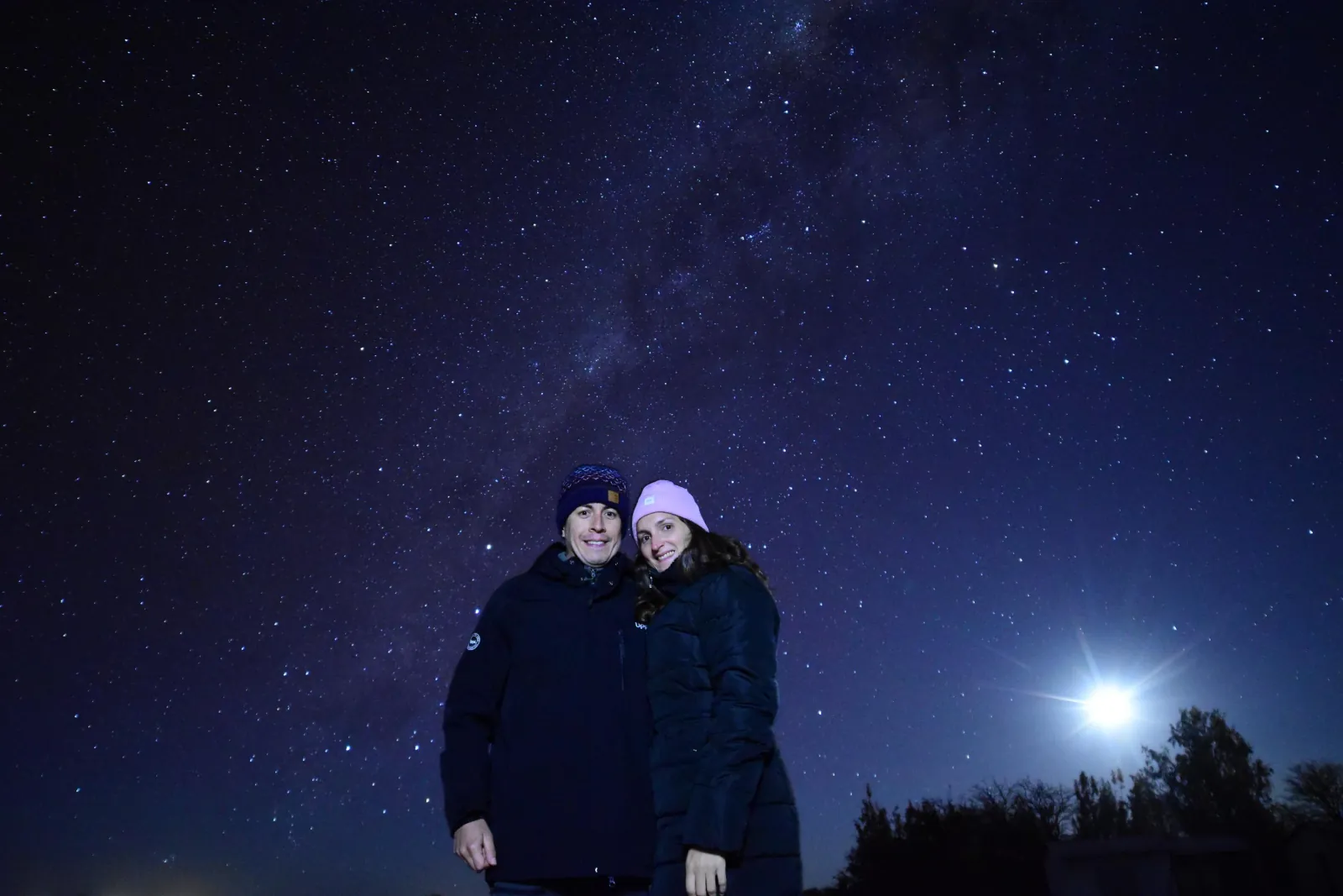 A couple in winter coats and woolly hats smiles under a star-filled sky with the moon glowing above dark trees