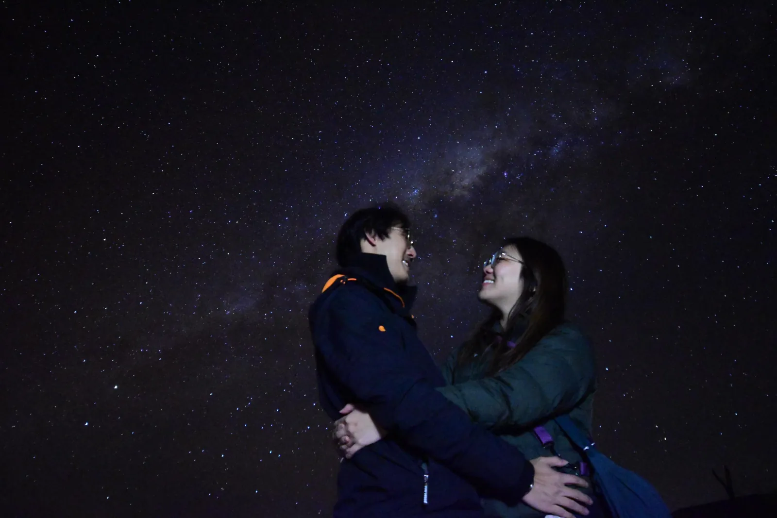 A laughing couple in dark jackets embraces under the Milky Way at the observation site outside San Pedro
