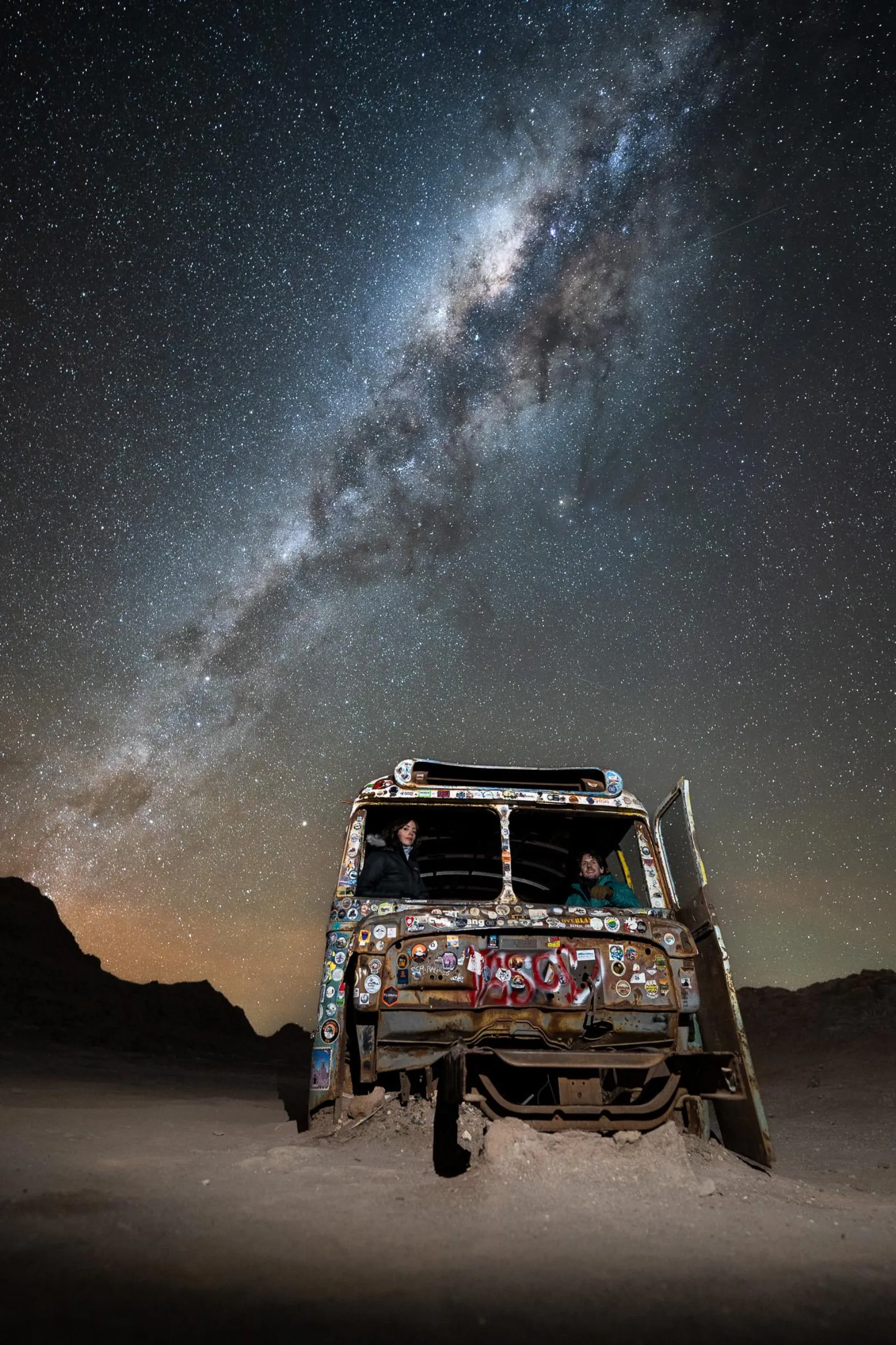 A colorfully painted old truck parked in the desert under the arching Milky Way, with dark rock ridges behind