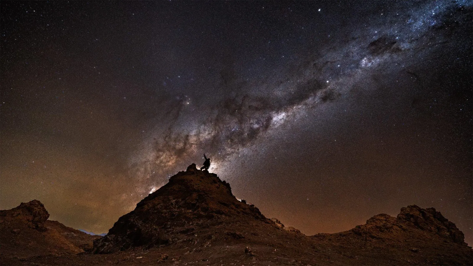 A lone figure with raised arms stands atop a rocky desert peak as the Milky Way core arcs overhead