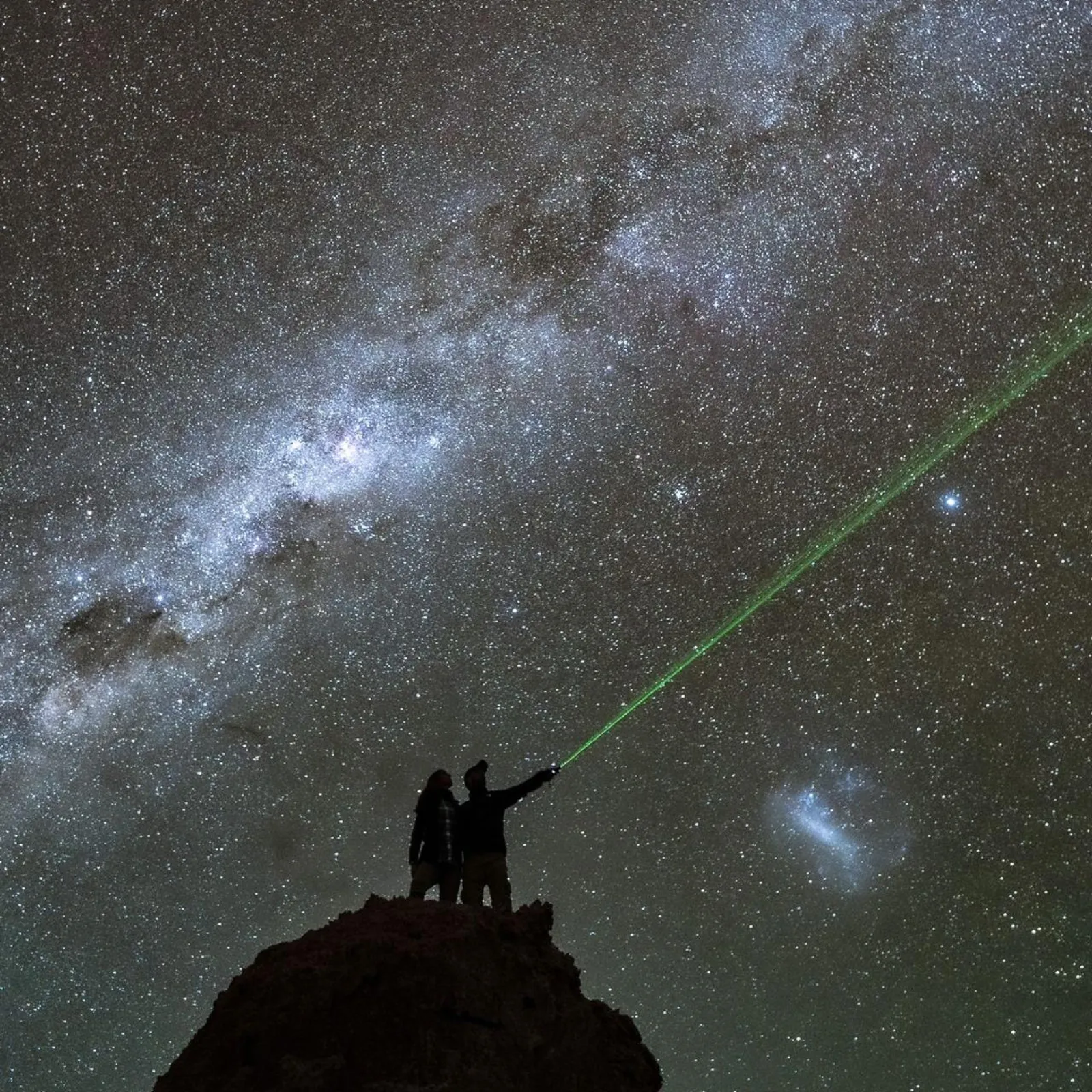 Two people on a rock outcrop aim a green laser at the sky beside the Milky Way and the Large Magellanic Cloud