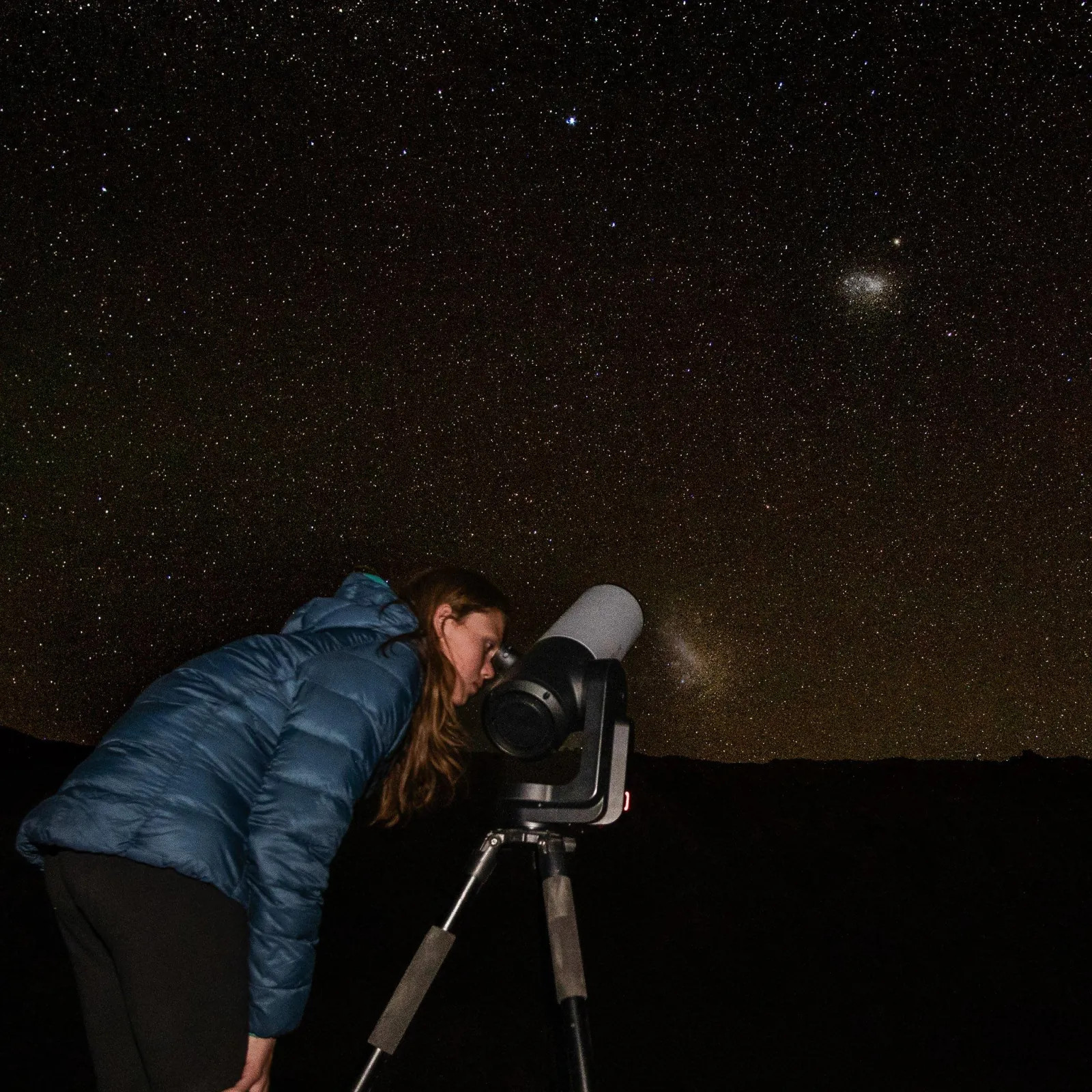 A guest in a blue puffer jacket looks into a white smart telescope on a tripod under a dark starry sky