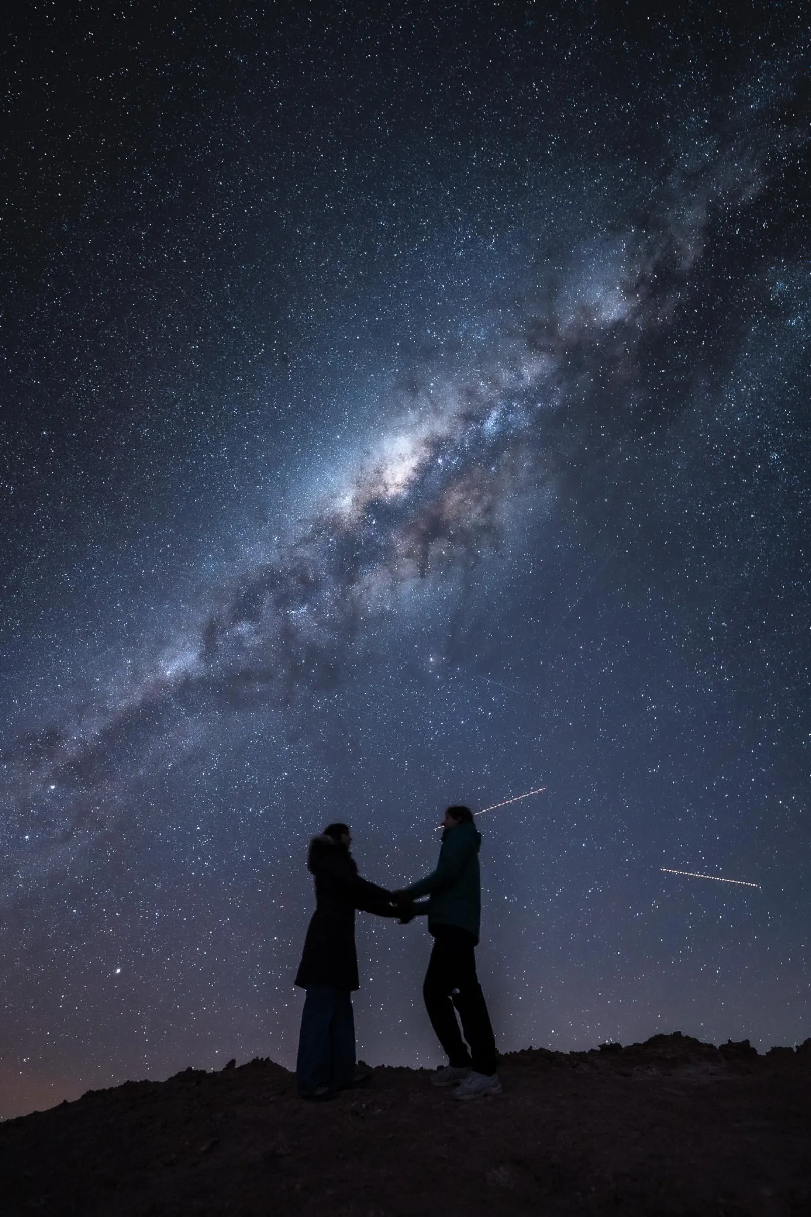 Two people stand facing each other on a desert ridge under the Milky Way as thin light streaks cross the sky