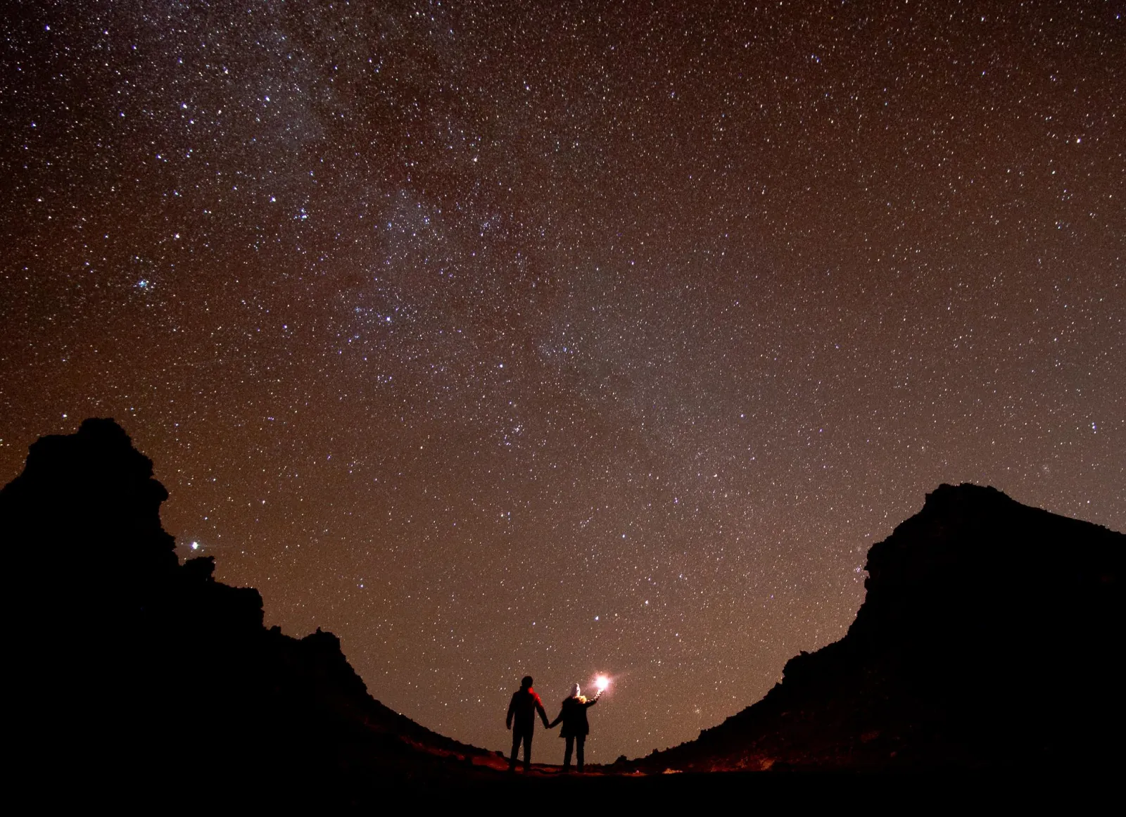 Two small figures holding a light stand in a rocky desert valley under a sky full of stars