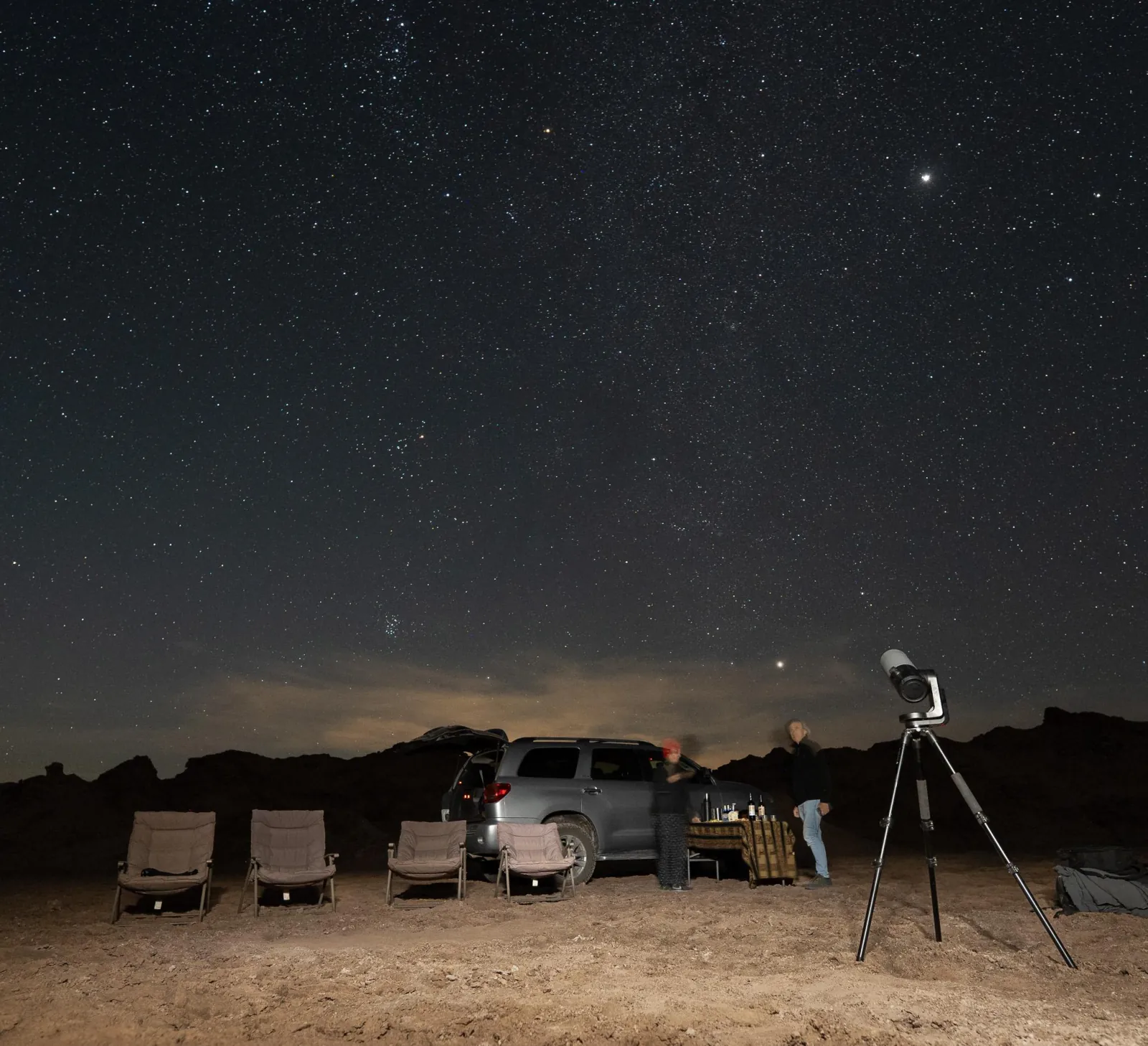 Folding chairs, a 4x4 and a smart telescope on a tripod set up on the desert floor under the stars