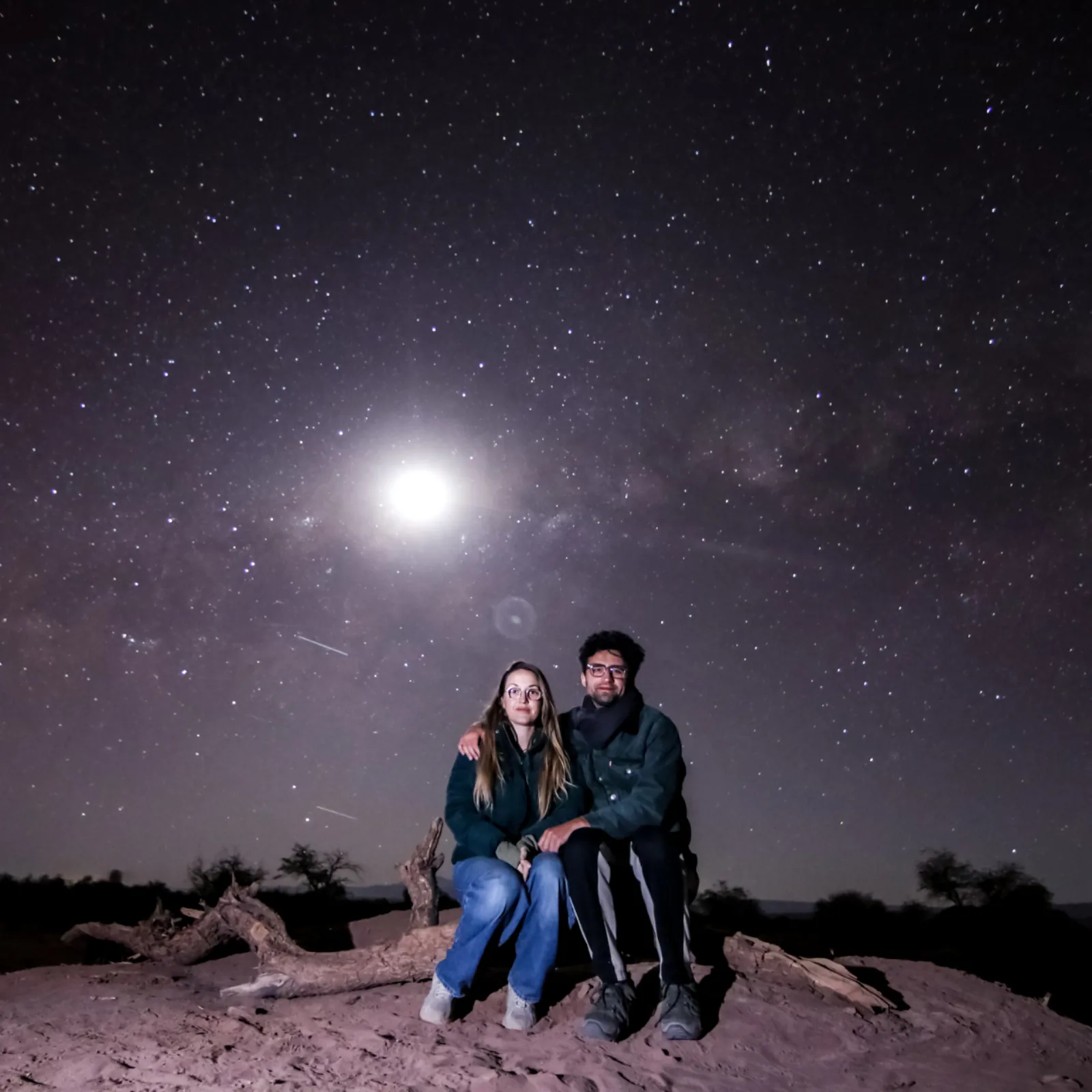 A couple sits on a log on the sand as a bright moon lights up the starry sky behind them