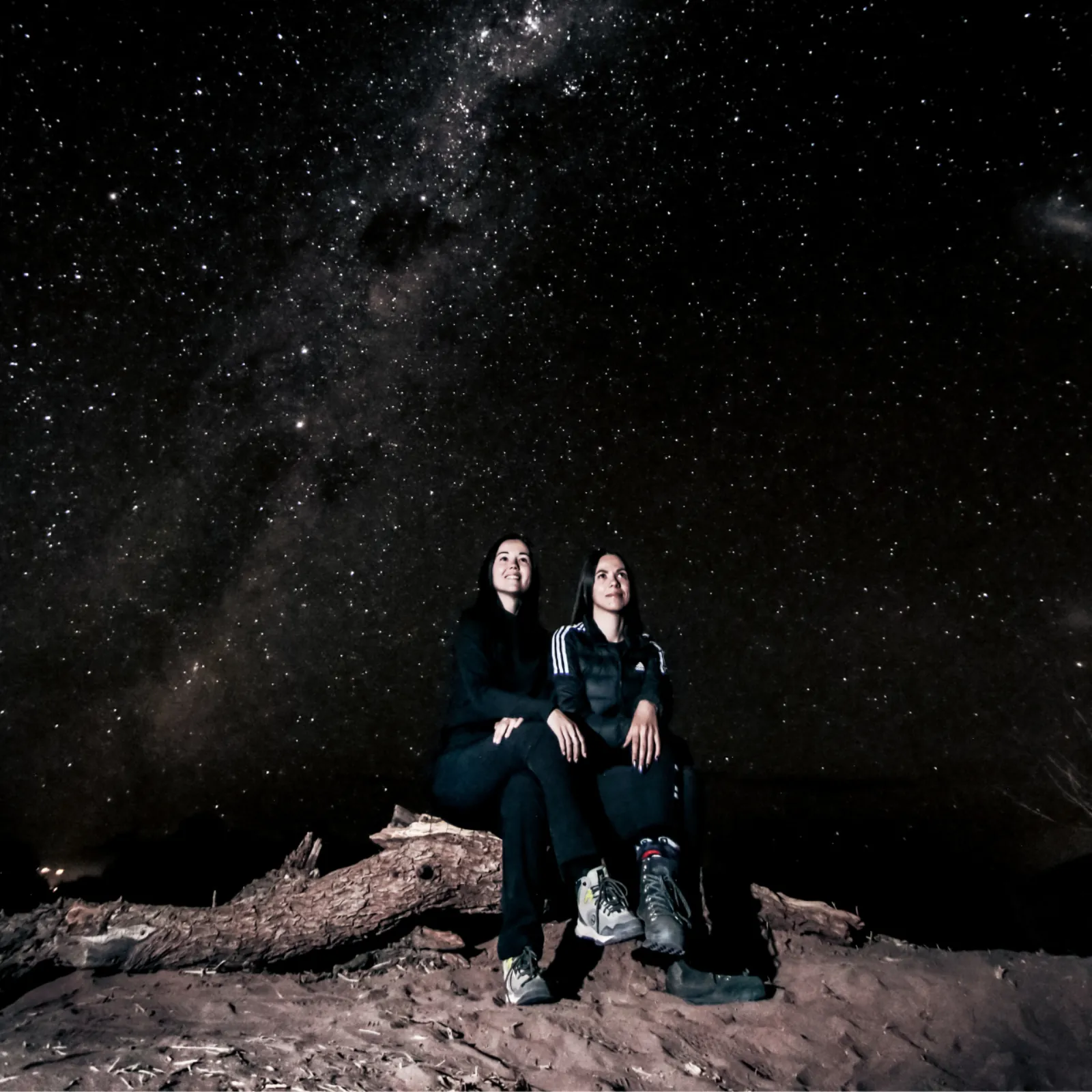 Two guests in dark jackets sit on a fallen log in the desert beneath the Milky Way