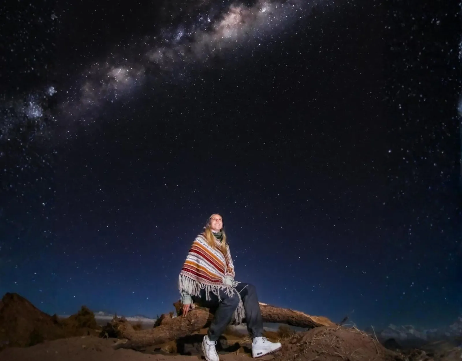 A guest in a striped Andean poncho sits on a desert log under the Milky Way with dark hills on the horizon