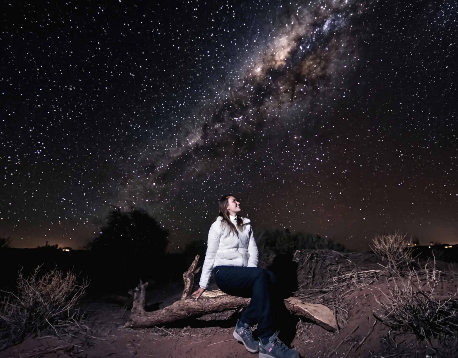 A woman in a white jacket sits on a fallen log among desert shrubs, looking up at the Milky Way