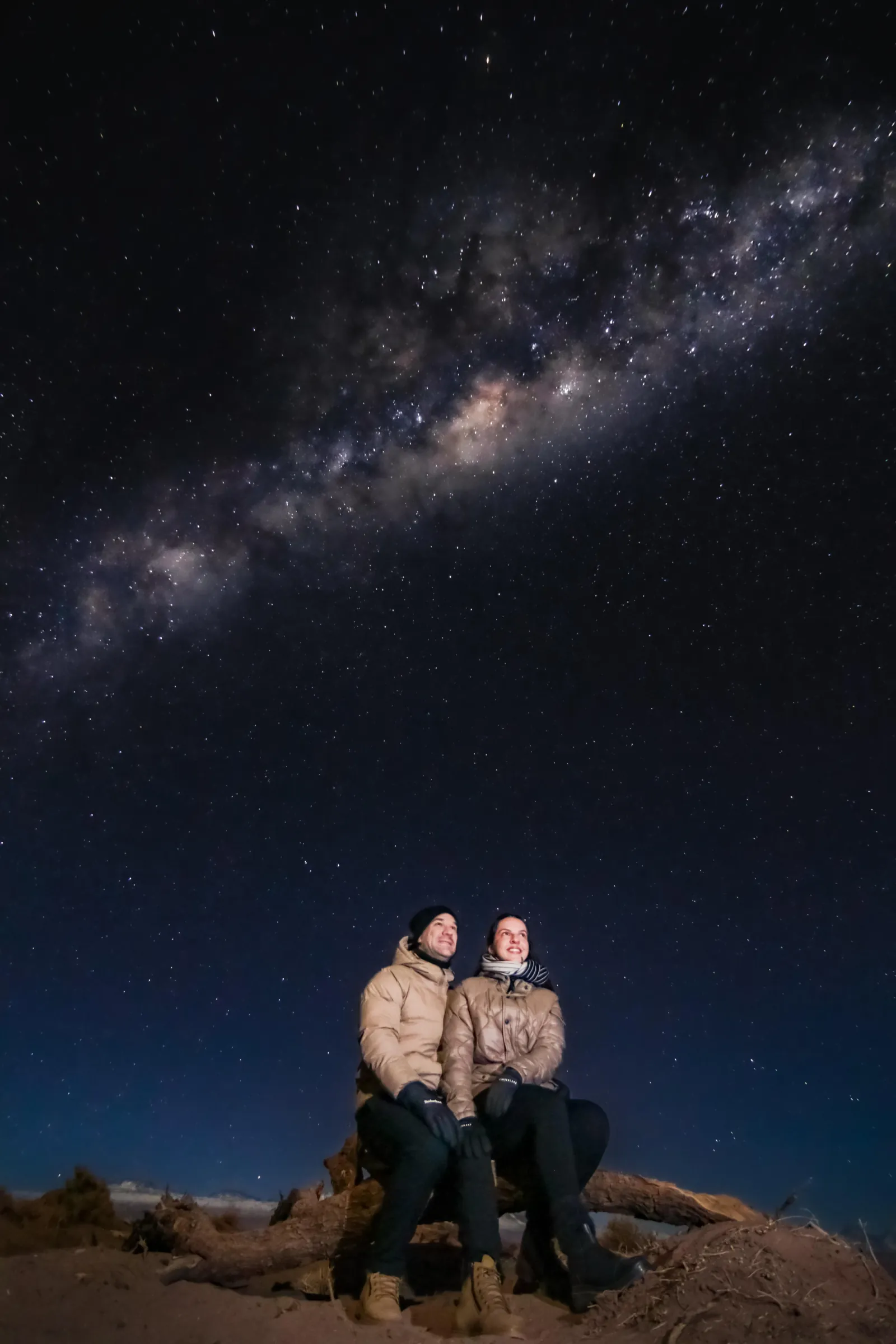 A couple in puffer jackets sits close together on a log under the Milky Way at a dark desert site