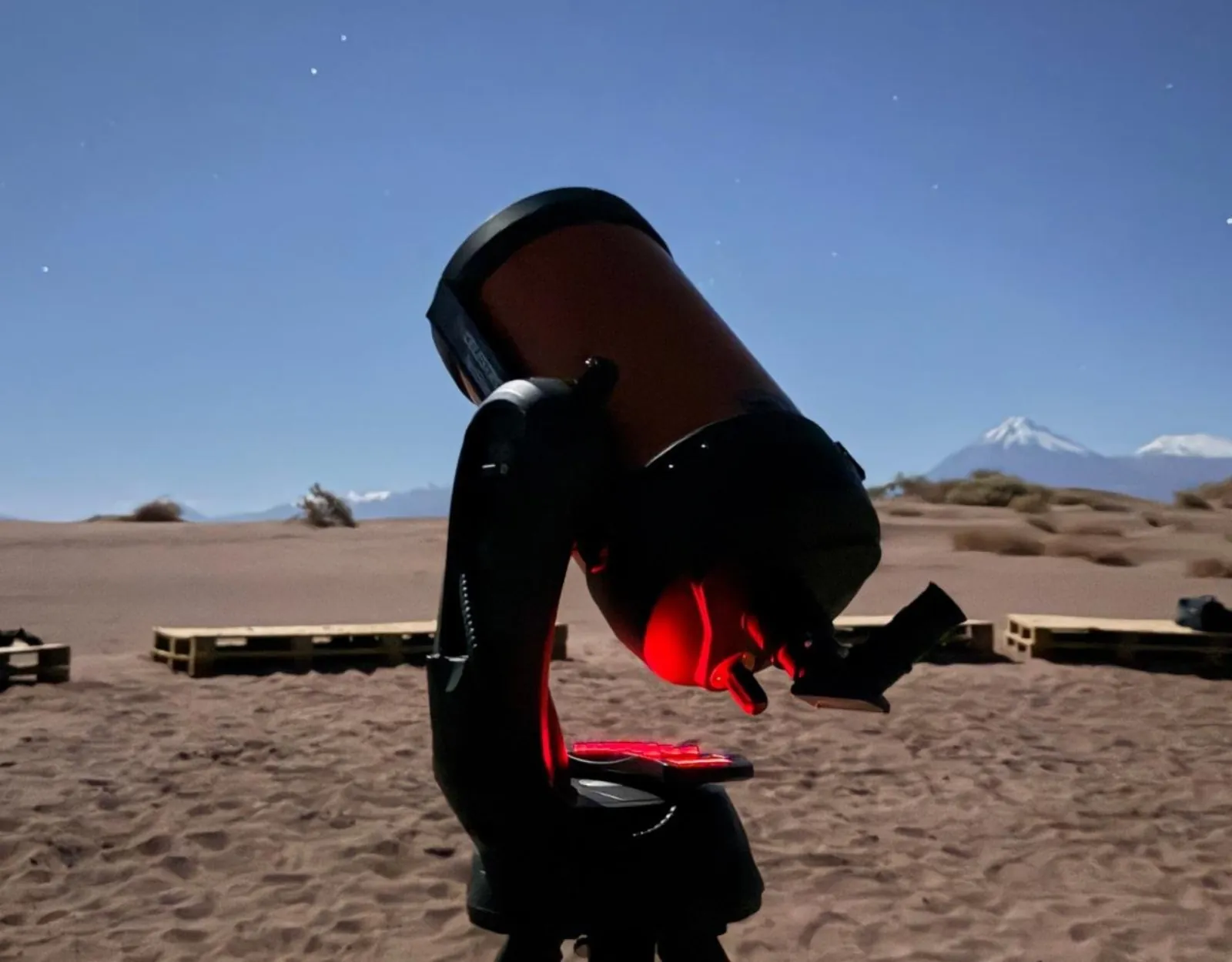 An orange-tubed telescope on sandy ground at dusk, with wooden benches behind and a snow-capped volcano on the horizon