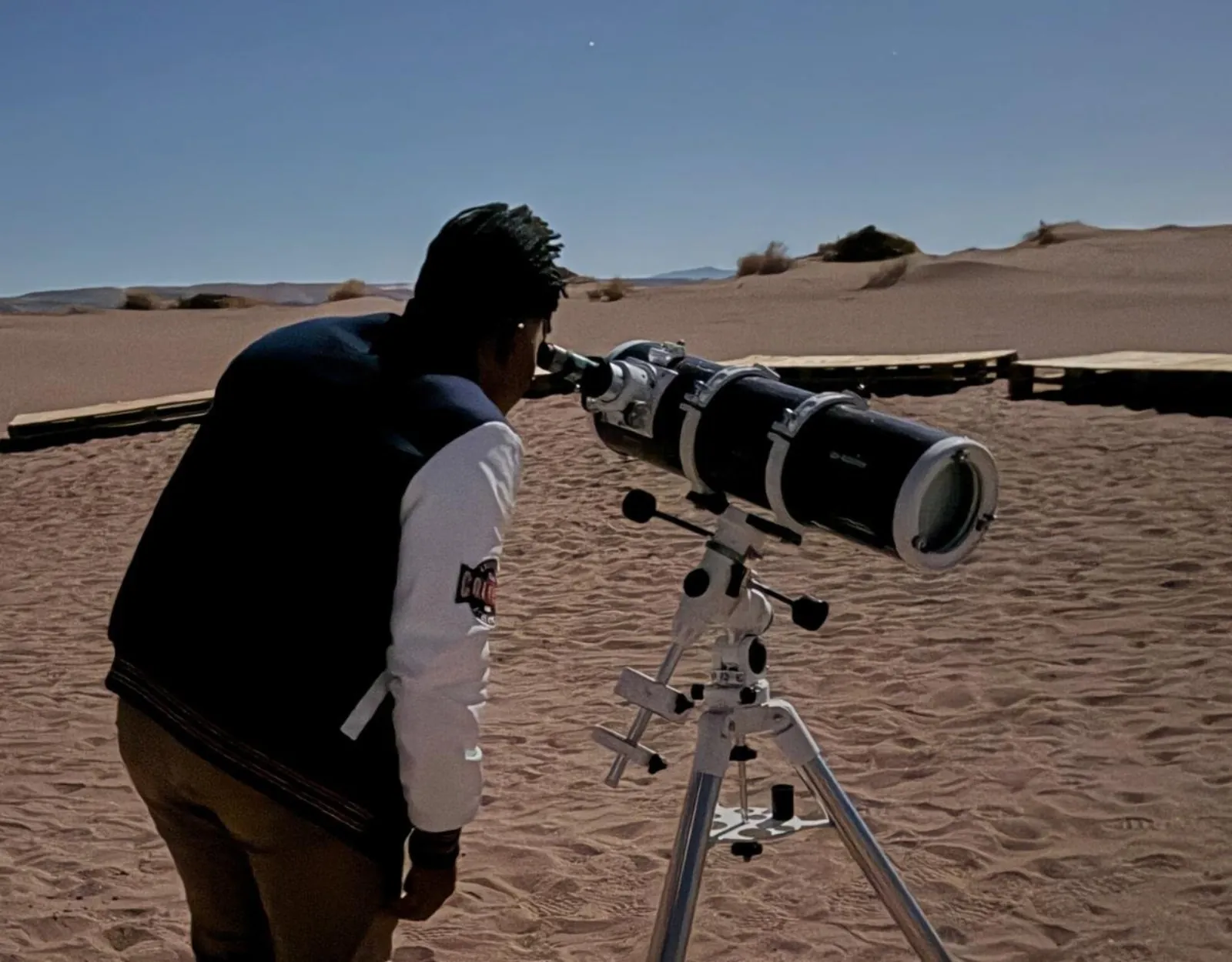 A guest bends to look through a white refractor telescope on a tripod set up on sand dunes at dusk