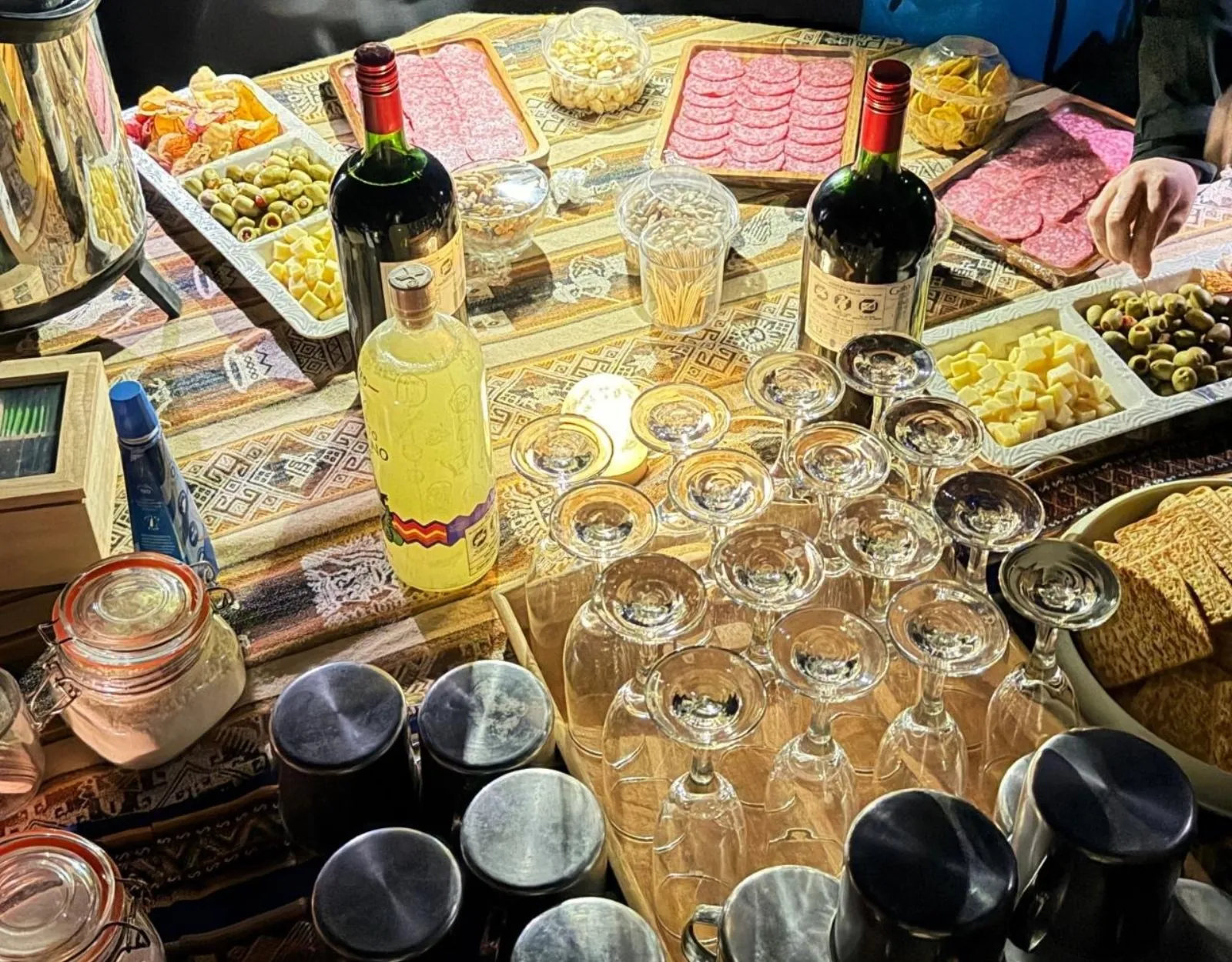 A tasting table laid with wine bottles, glasses, cheese, olives, salami and hot drink flasks on an Andean textile