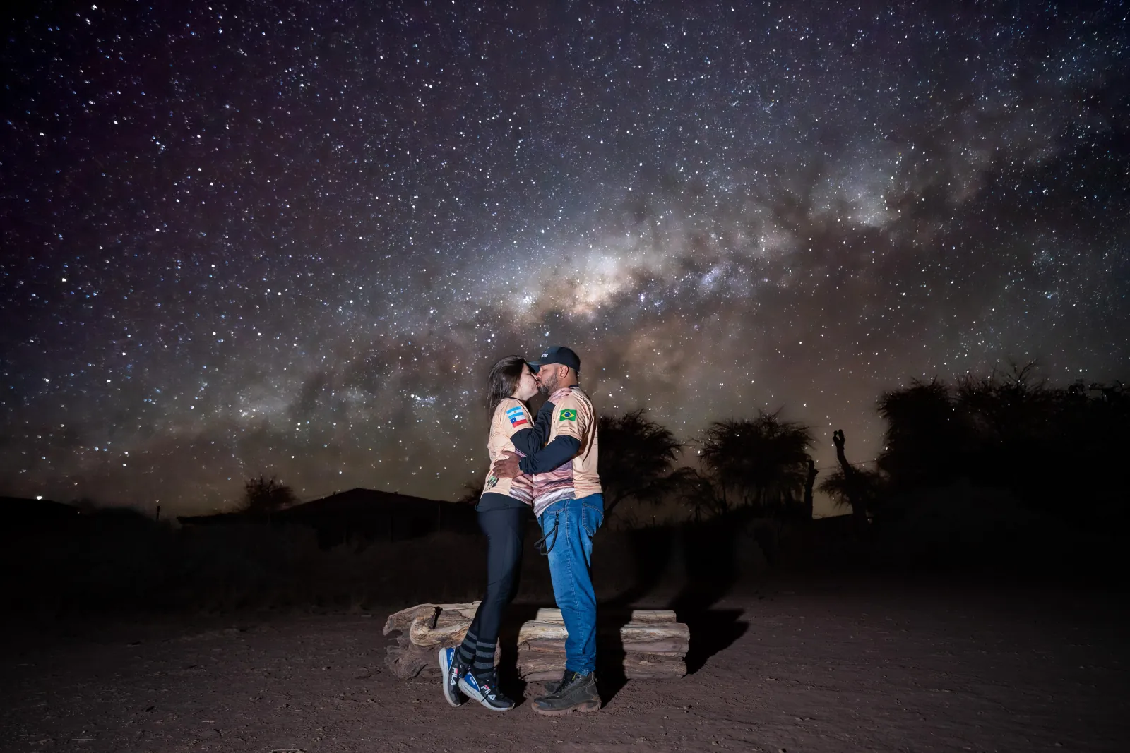 A couple kisses beside a log bench in the desert under the bright core of the Milky Way