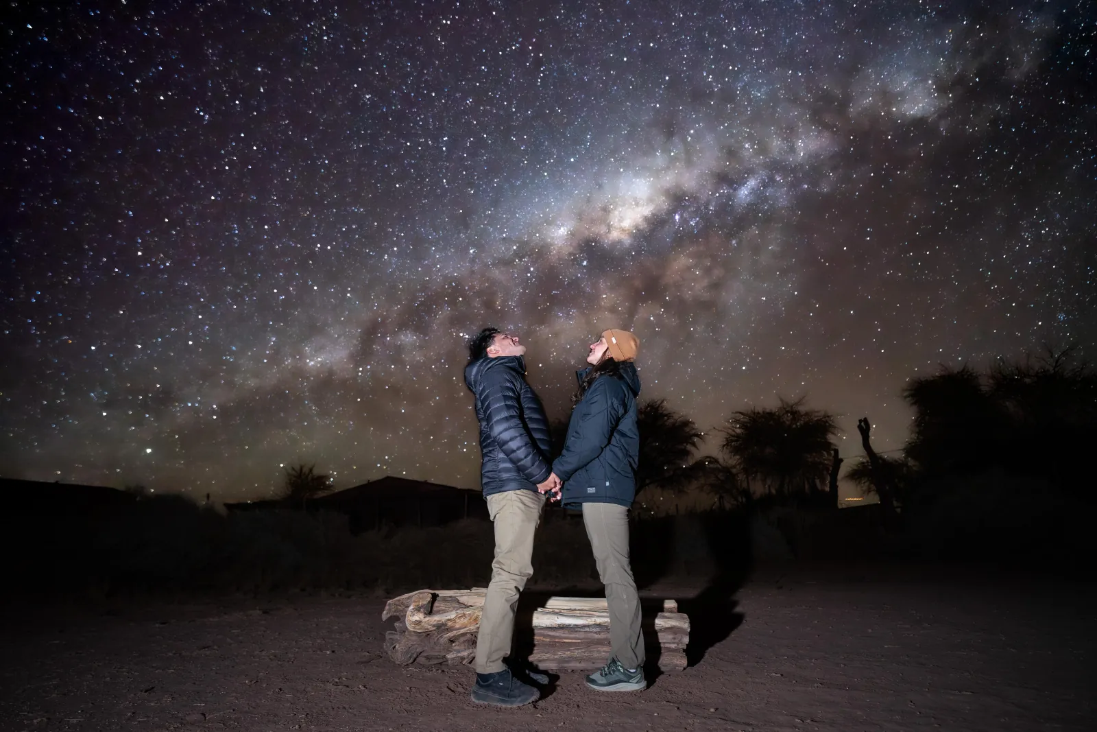 A couple in down jackets holds hands and looks up at the Milky Way core above the desert site