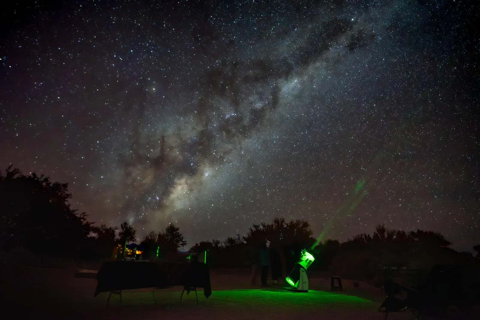 A green laser and green-lit telescopes at the observation site under the Milky Way, with tables set nearby