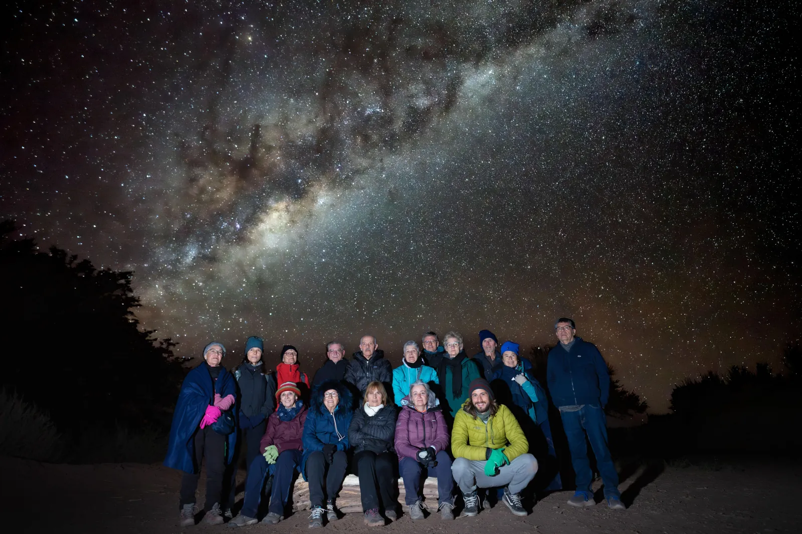 A group of about fifteen guests in warm jackets poses under the arching Milky Way at the desert site