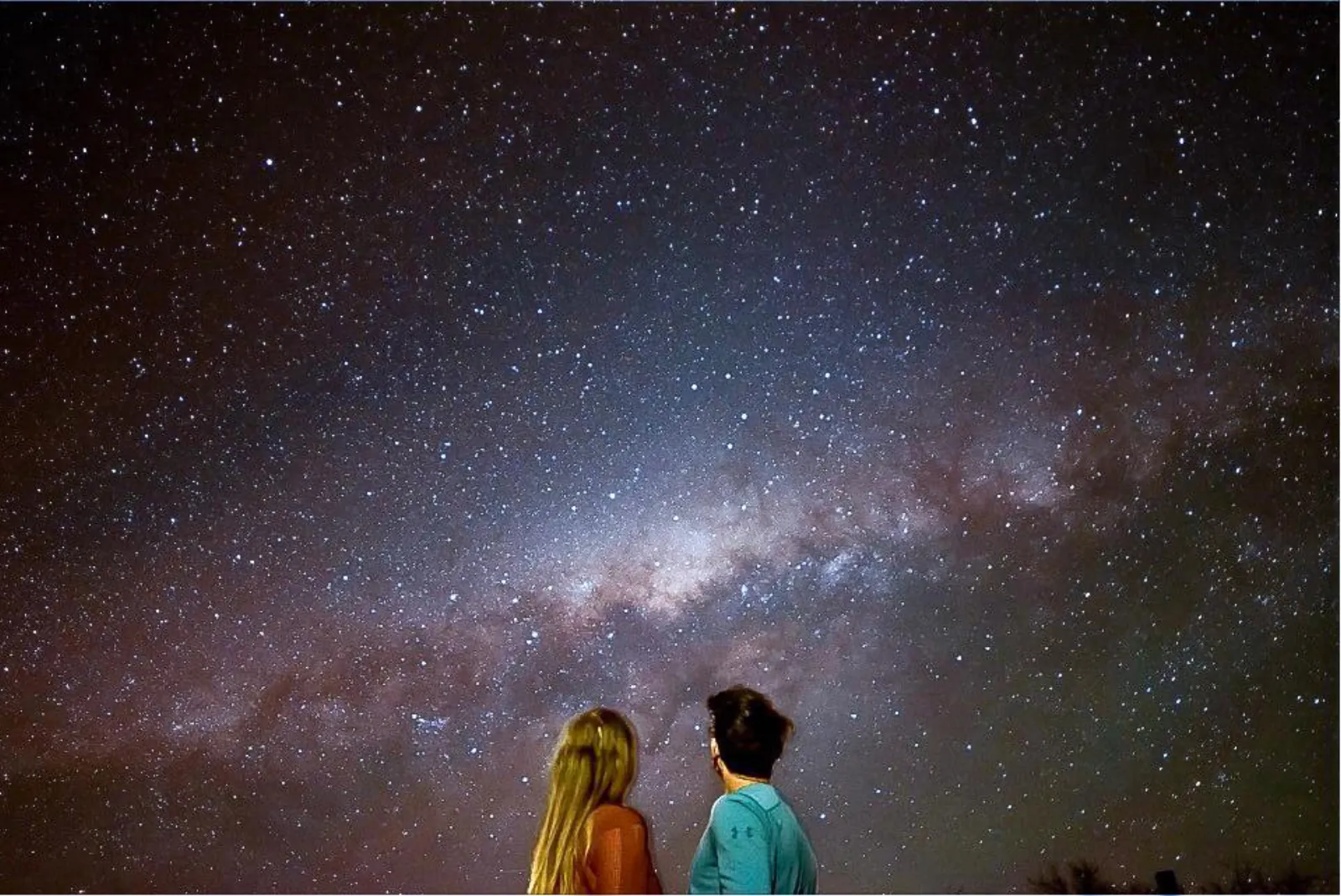Two guests seen from behind look up at the glowing band of the Milky Way across a star-filled desert sky