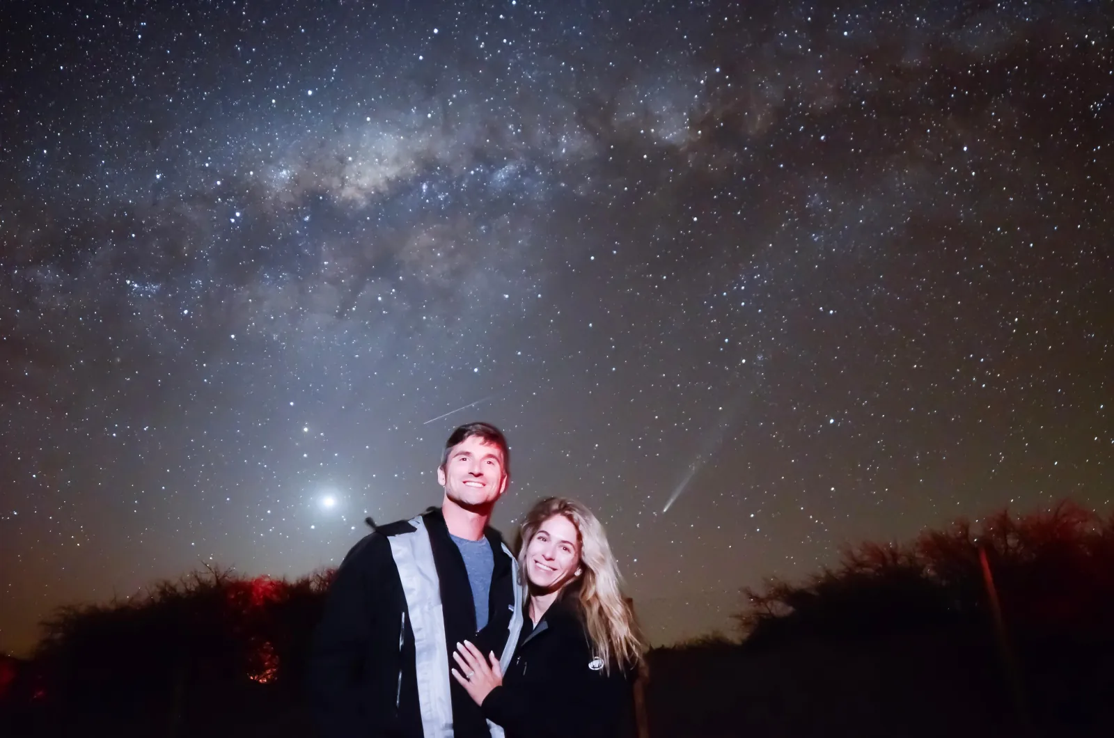 A smiling couple poses at night beside a bright planet low in the sky and a faint comet tail above red-lit shrubs