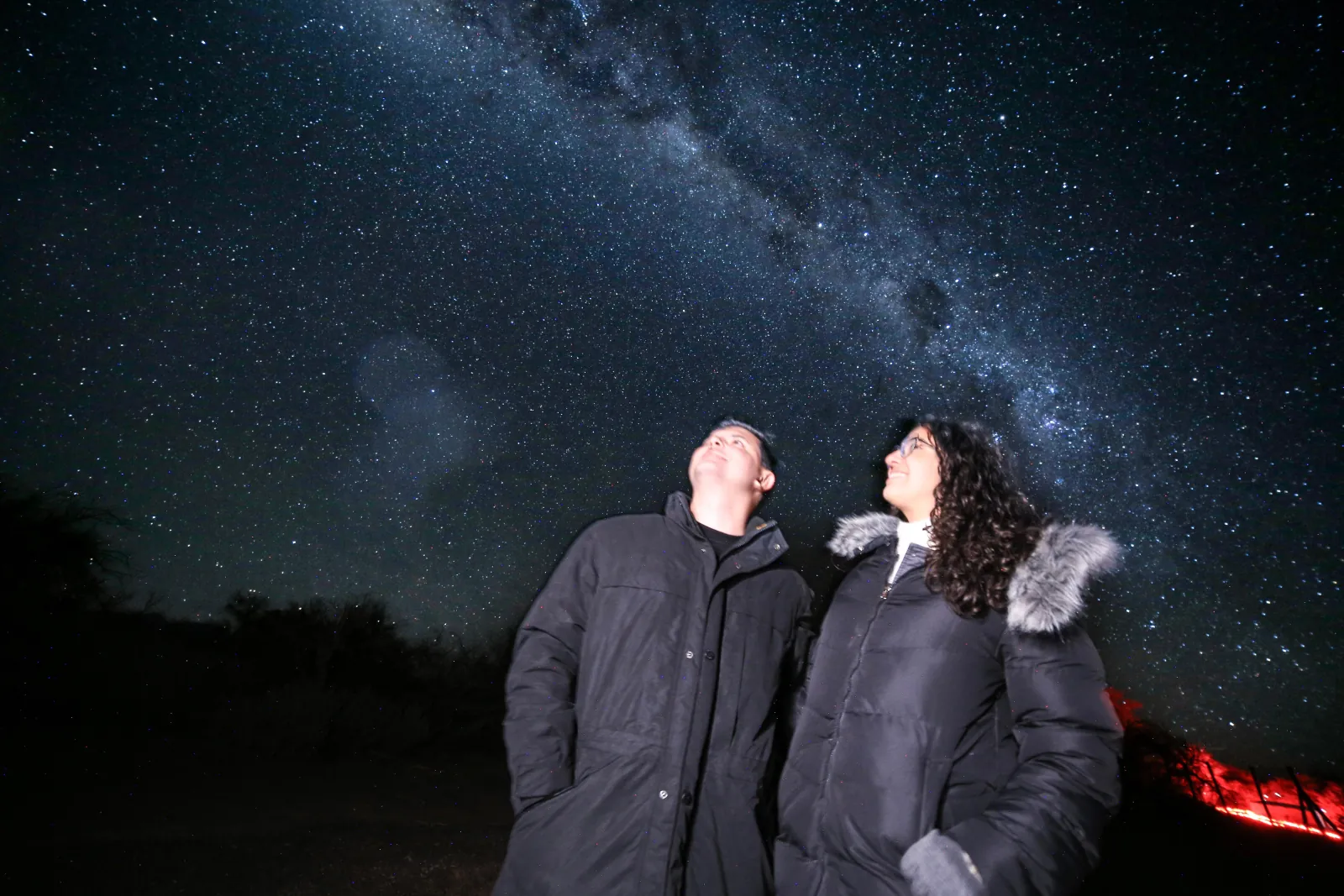 Two guests in heavy winter coats gaze up at the Milky Way as a red light glows on the ground behind them