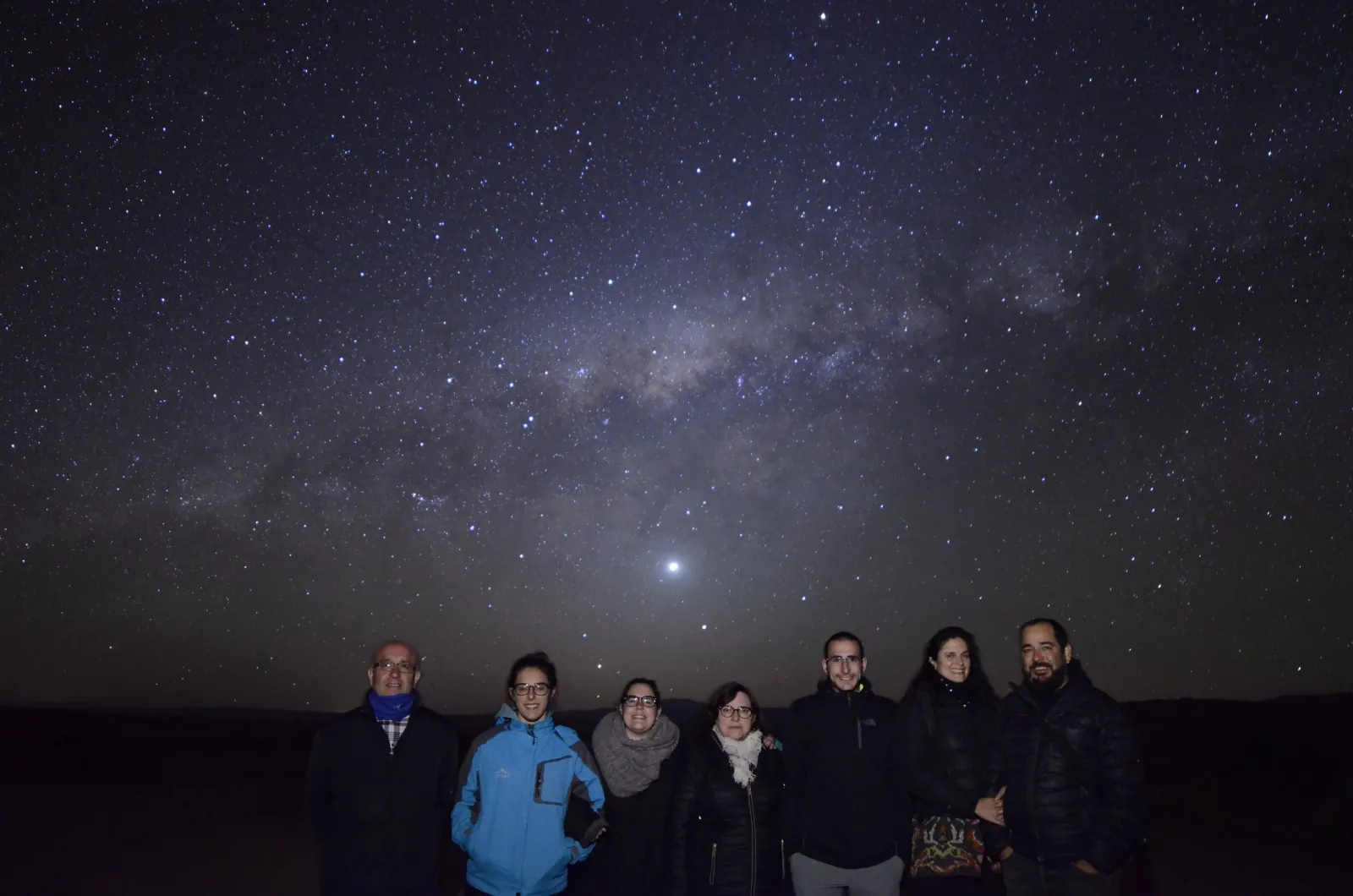 Seven guests in coats stand in a row on dark open ground under a starry sky with a bright planet above