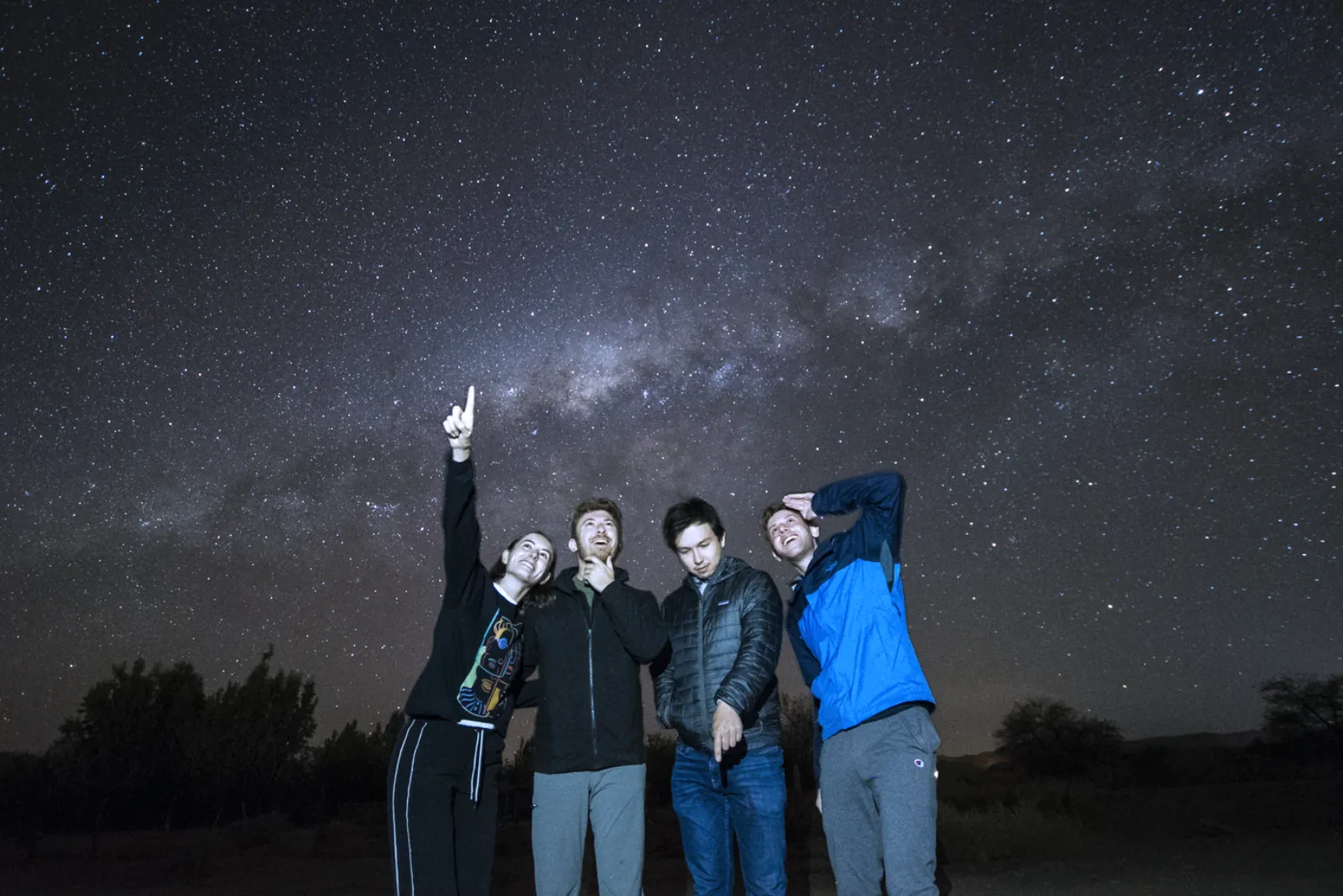 Four guests in jackets point and gaze up at the Milky Way arching over dark desert shrubs outside San Pedro de Atacama