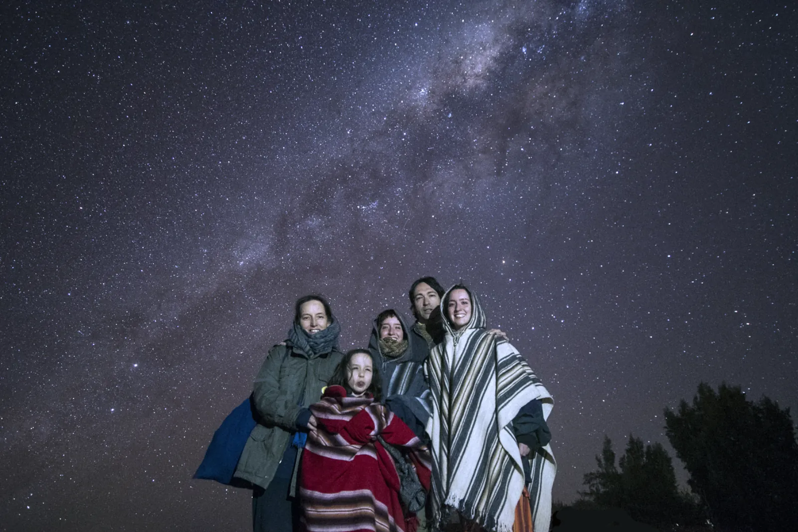 A family wrapped in striped Andean blankets and ponchos poses under a star-filled sky and the band of the Milky Way