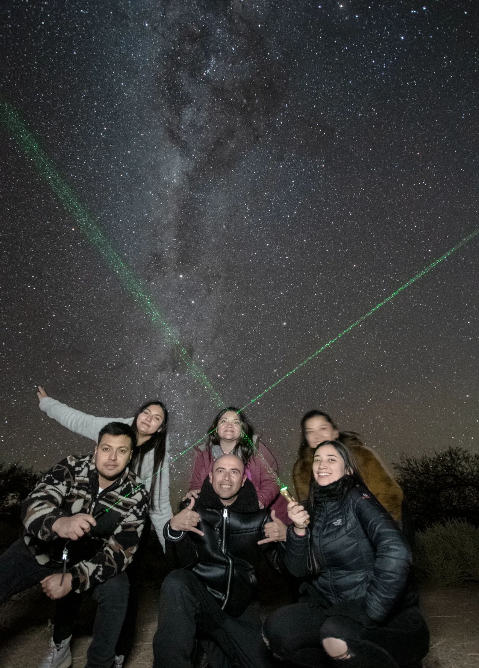 A smiling group crouches together as two green laser beams point out stars and the Milky Way above them