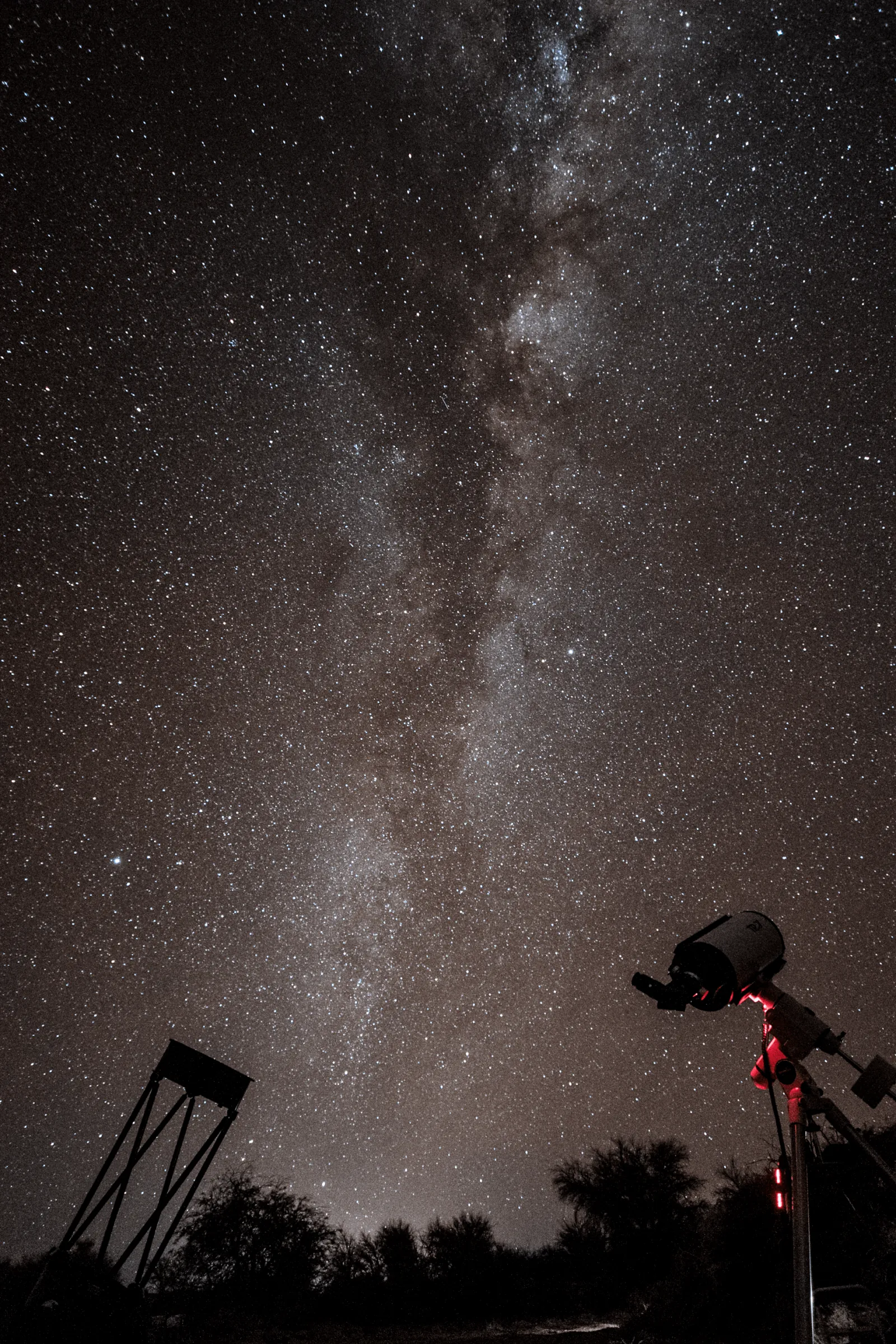 A telescope on its mount stands beside a tall frame structure under the vertical band of the Milky Way