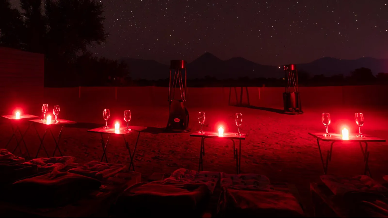 Red-lit stargazing area with candlelit tables, wine glasses, floor cushions and two large telescopes before dark hills