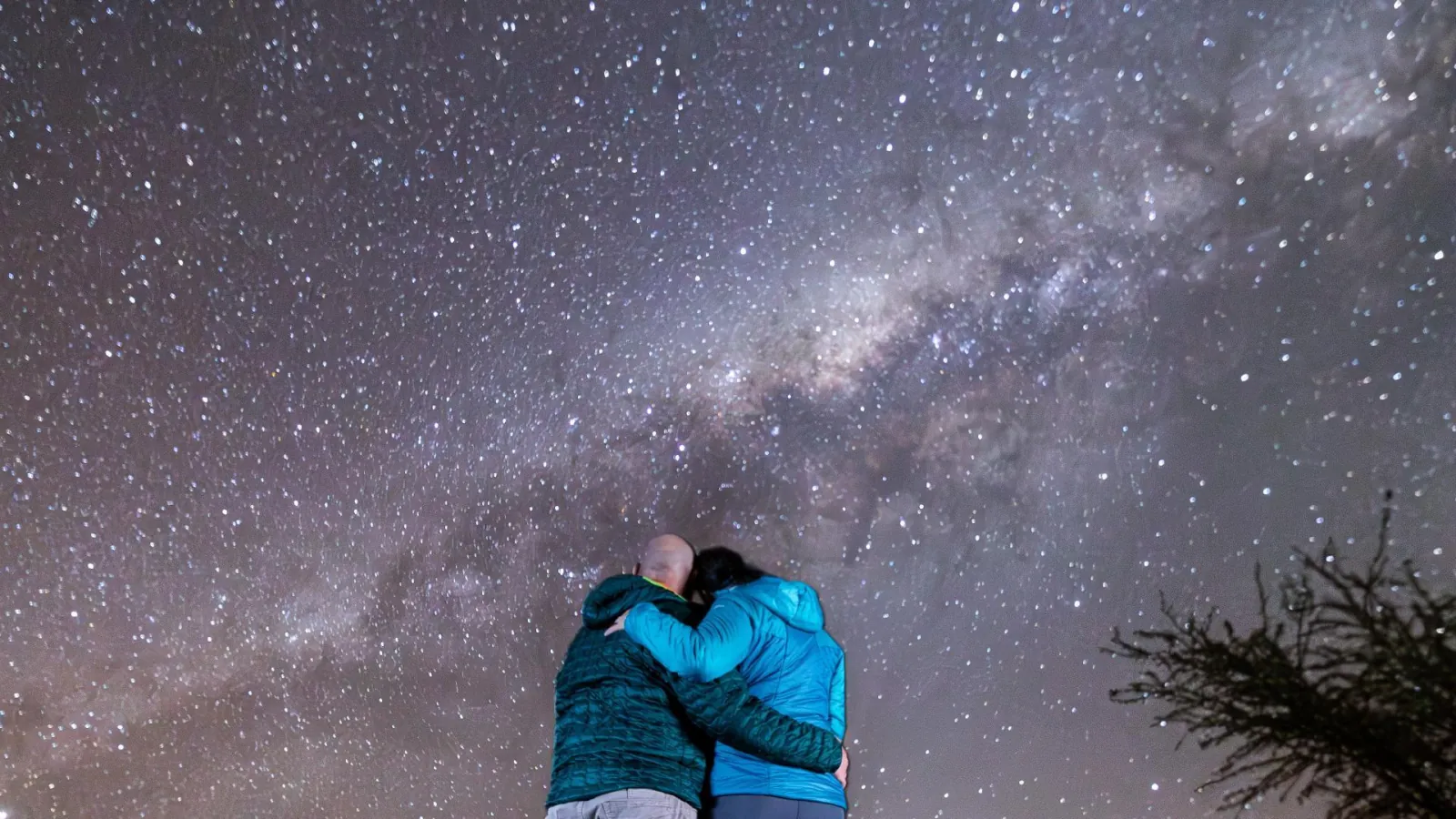 A couple in blue jackets hugs with their backs to the camera, facing the Milky Way over the Atacama Desert