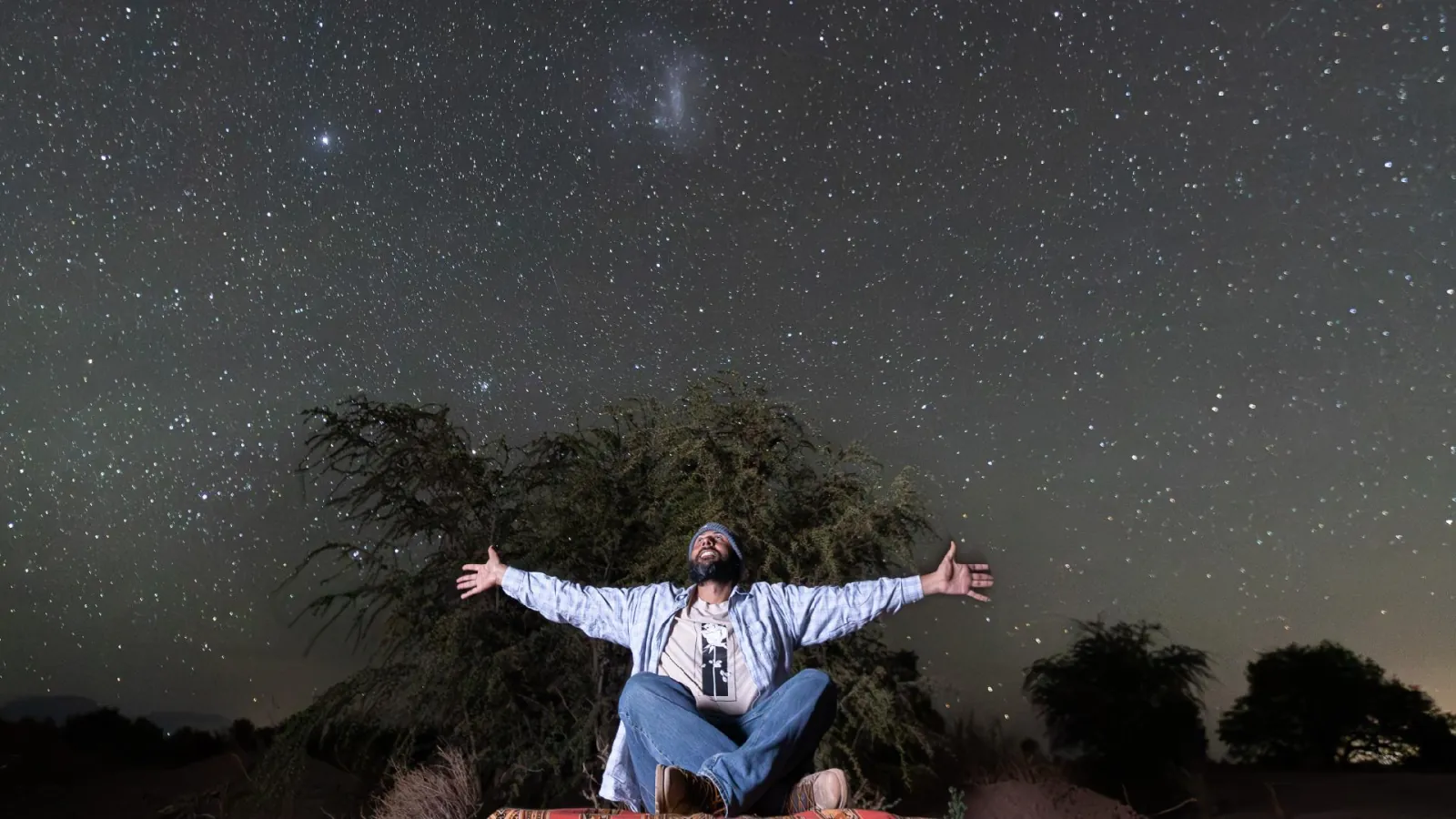 A guest sits cross-legged with arms spread wide below the Magellanic Clouds and a sky full of stars