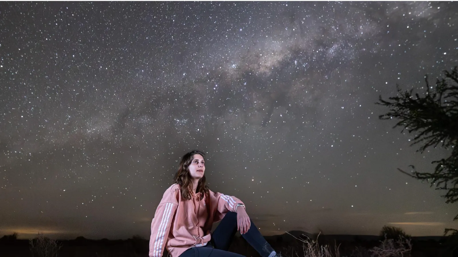 A young woman in a pink jacket sits on the desert floor gazing up at the Milky Way near San Pedro