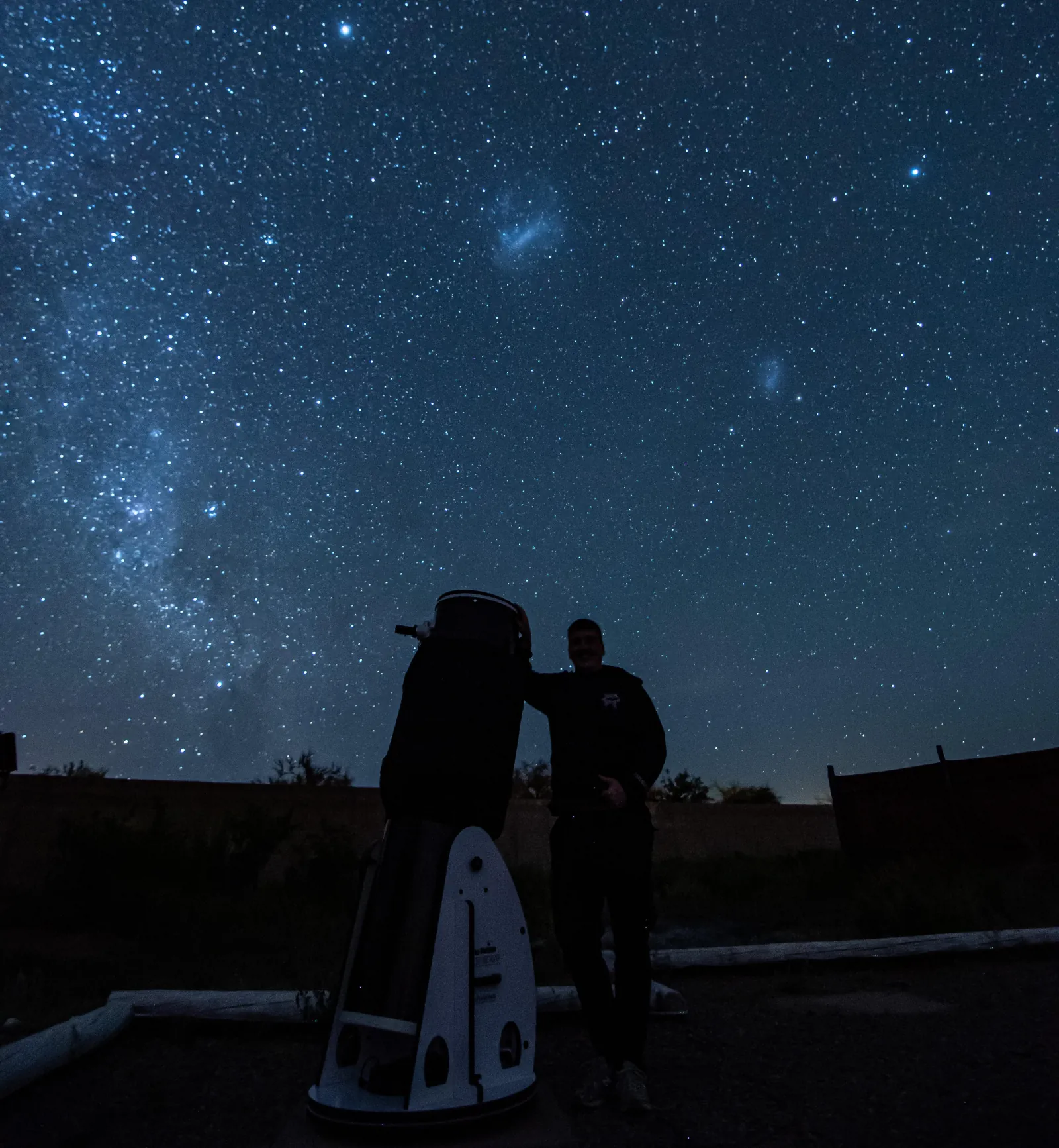 A person stands beside a large white Dobsonian telescope under the Milky Way and the Magellanic Clouds