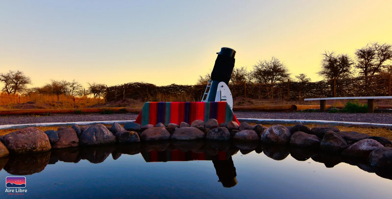 A large telescope and a striped Andean blanket reflected in the still water pool at sunset at the observation site