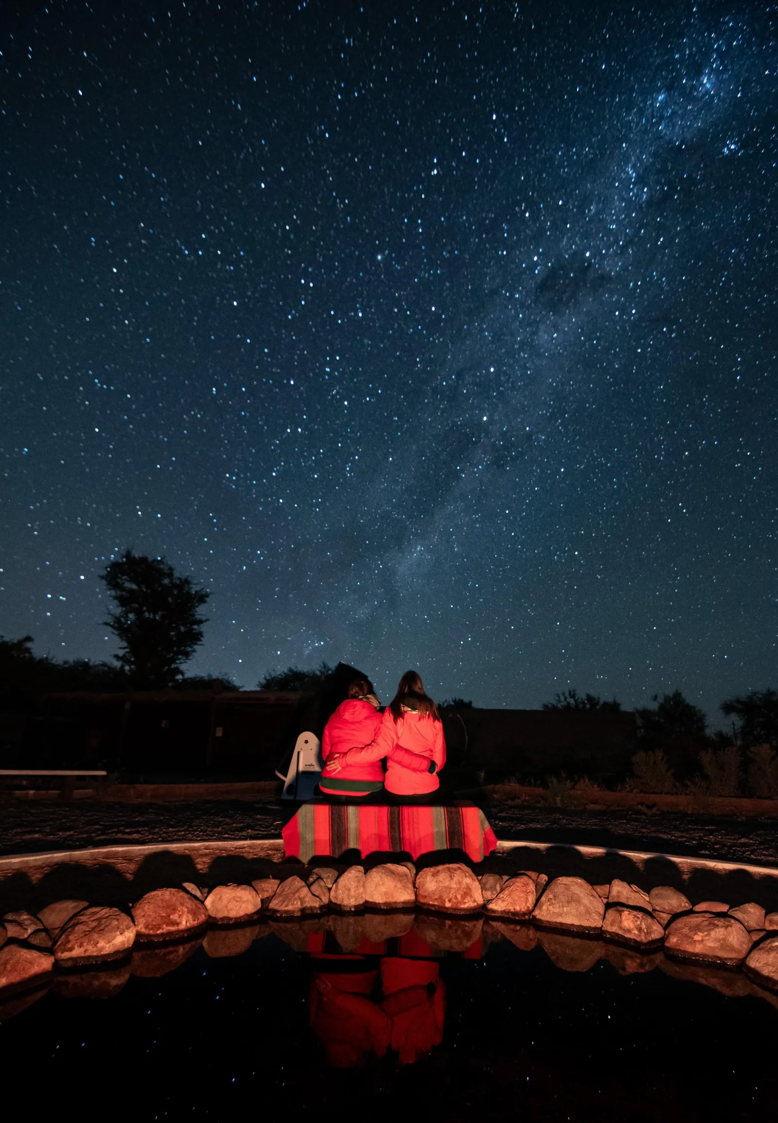 Two guests sit on a striped blanket by the water pool, looking up at the Milky Way in a starry sky