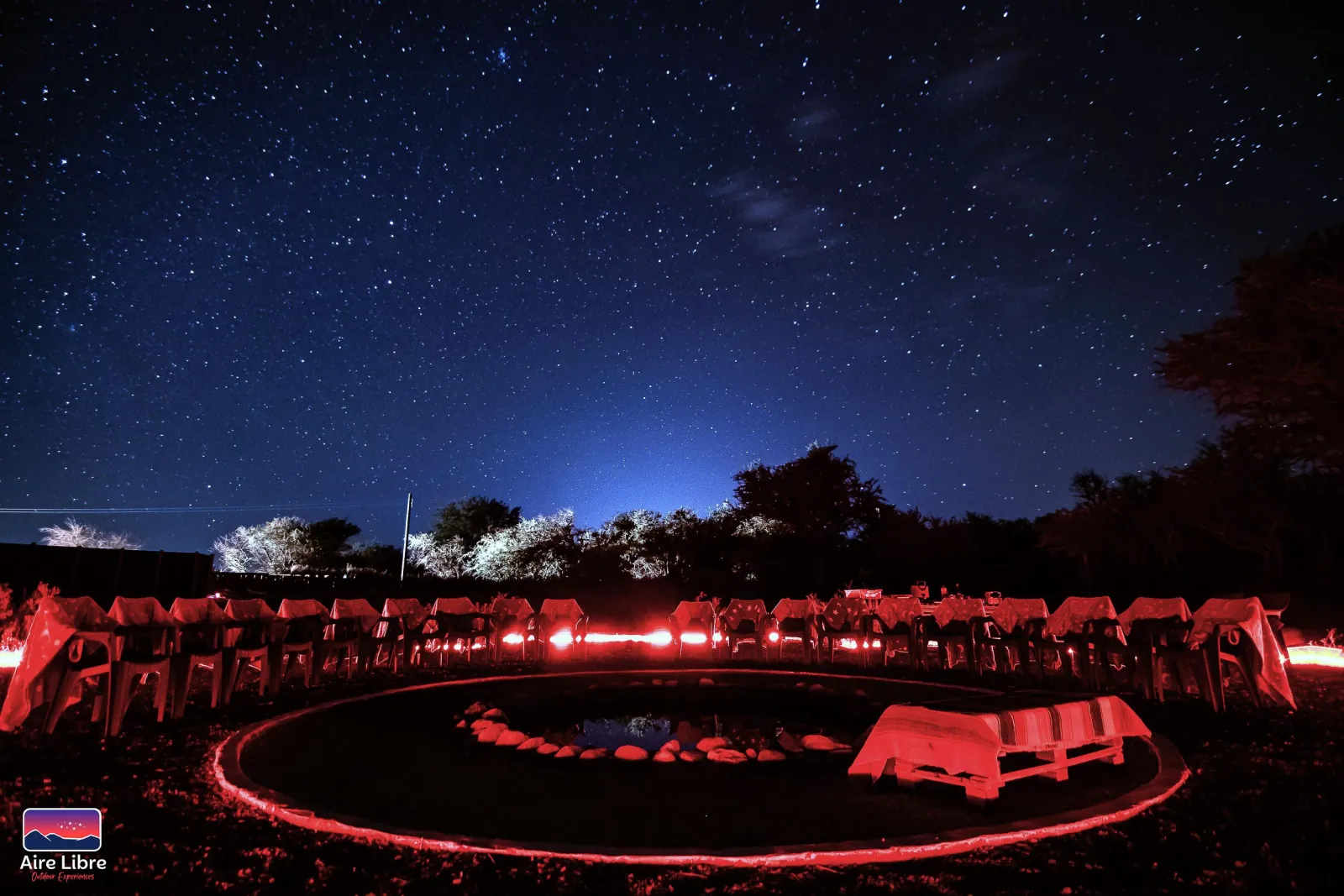 A ring of chairs and the stone-edged water pool glow under red light beneath a sky full of stars