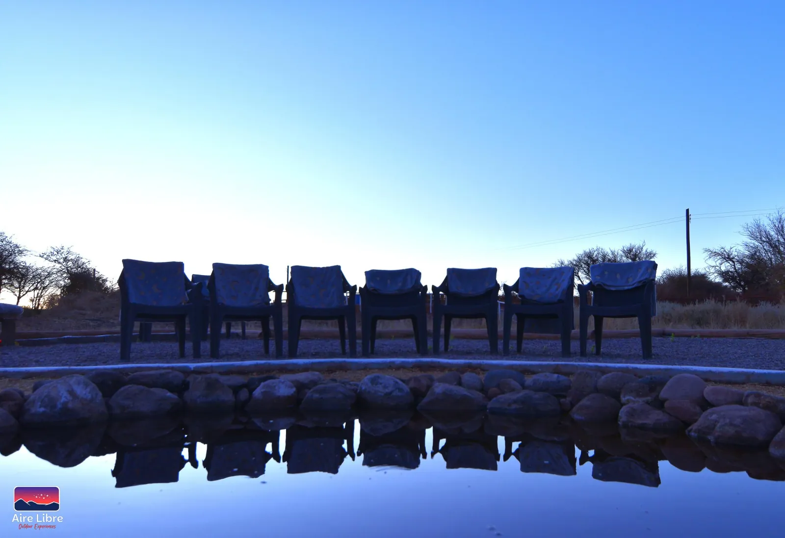 A row of chairs draped in blankets reflected in the water pool under a clear blue twilight sky