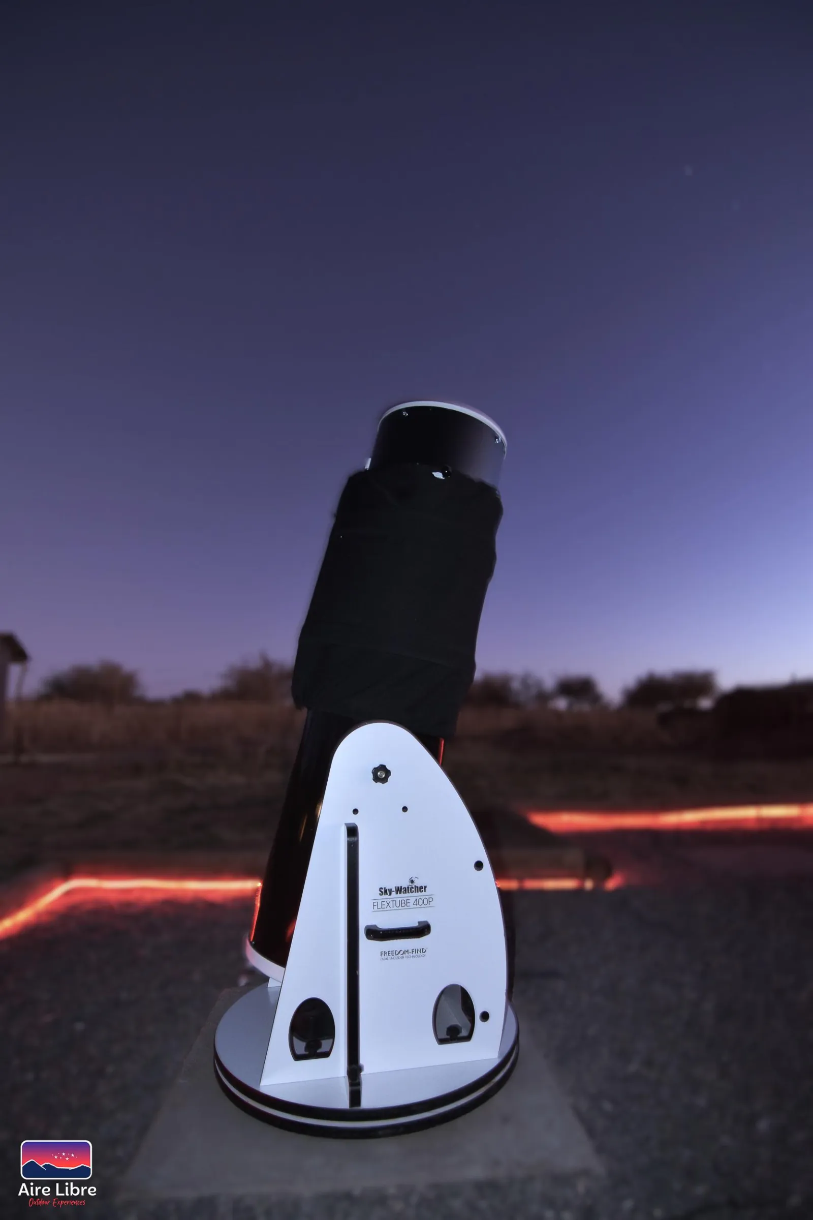 A tall white Dobsonian telescope stands on the observation area at dusk, with a red-lit path behind it